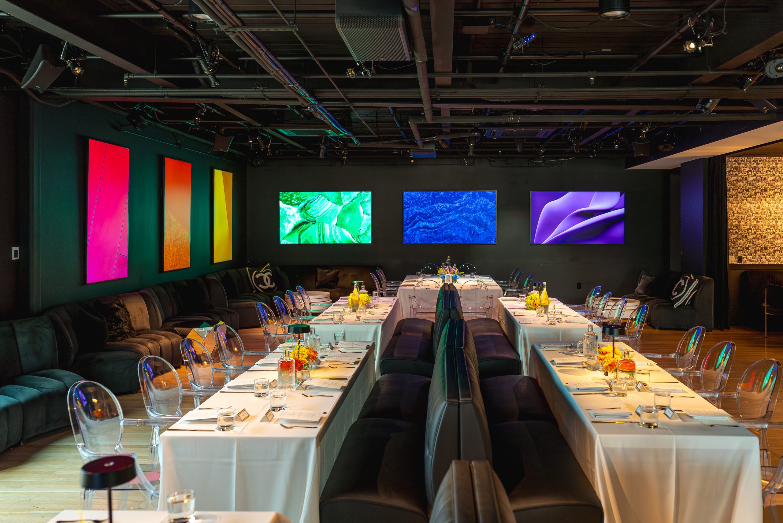 Event space set for a Pride dinner with long white-clothed tables and glowing colorful art screens on the walls