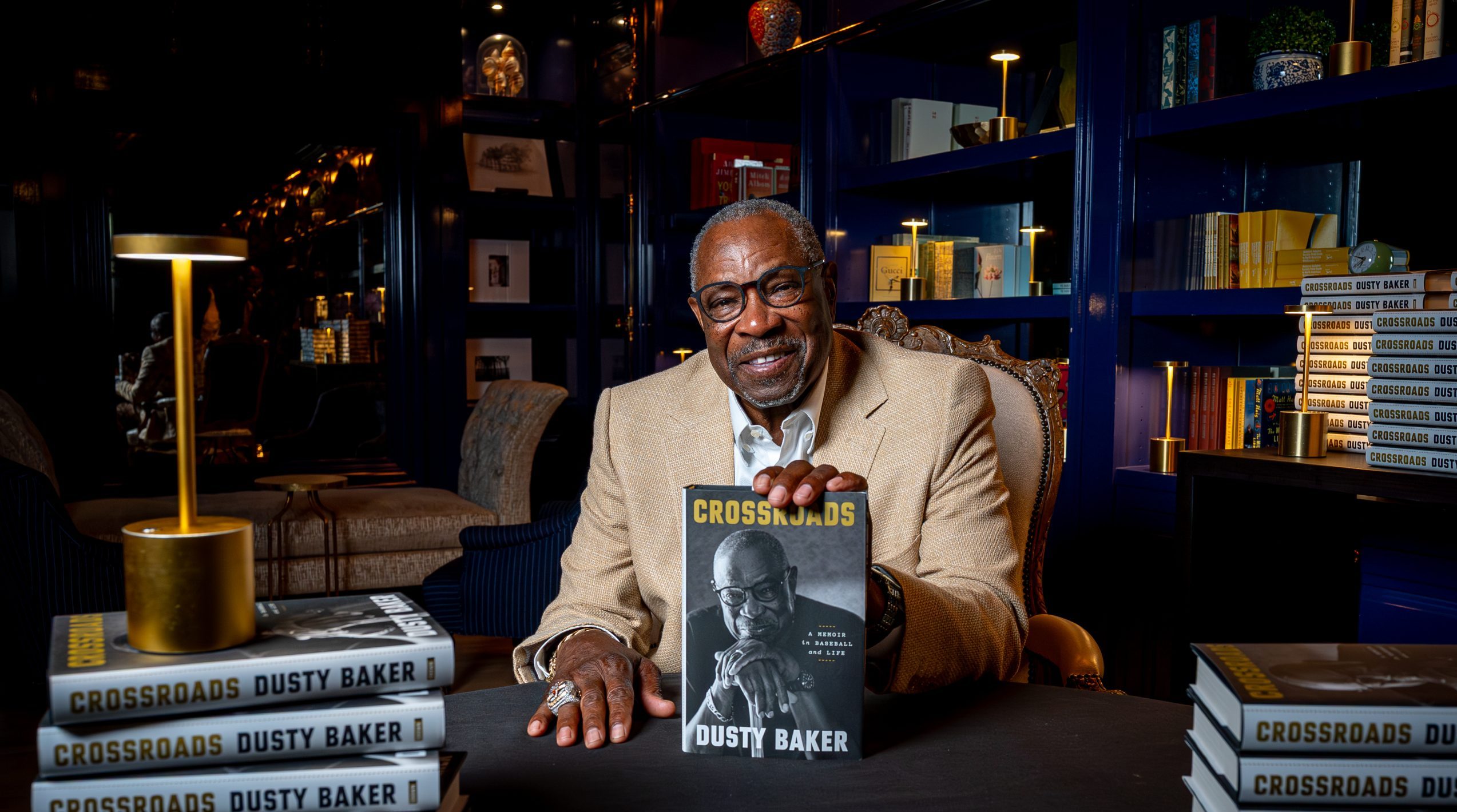 Dusty Baker smiling at a signing table in The Library, holding his memoir Crossroads beside stacks of the book