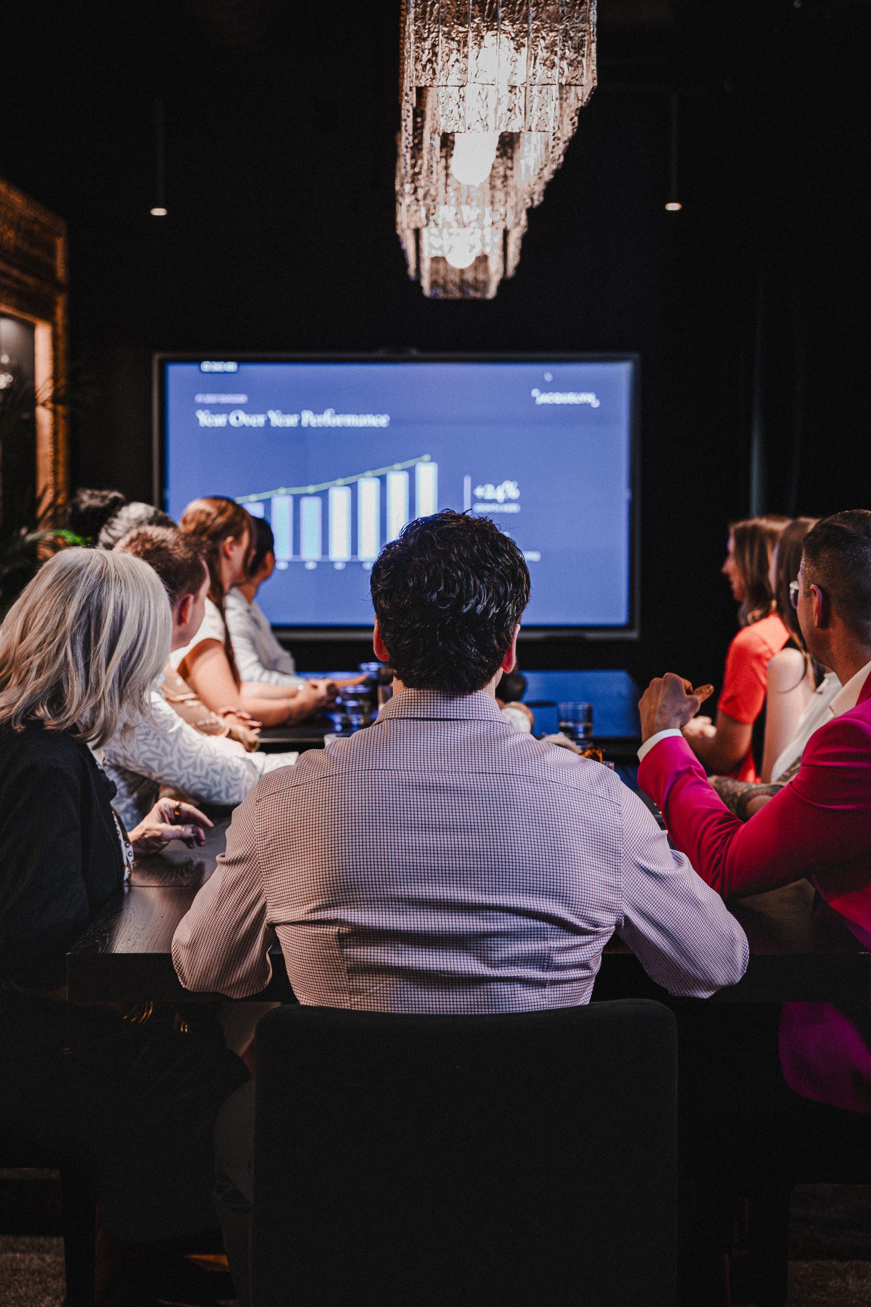 Team in the Boardroom watches a year-over-year performance chart on a large screen under a crystal chandelier
