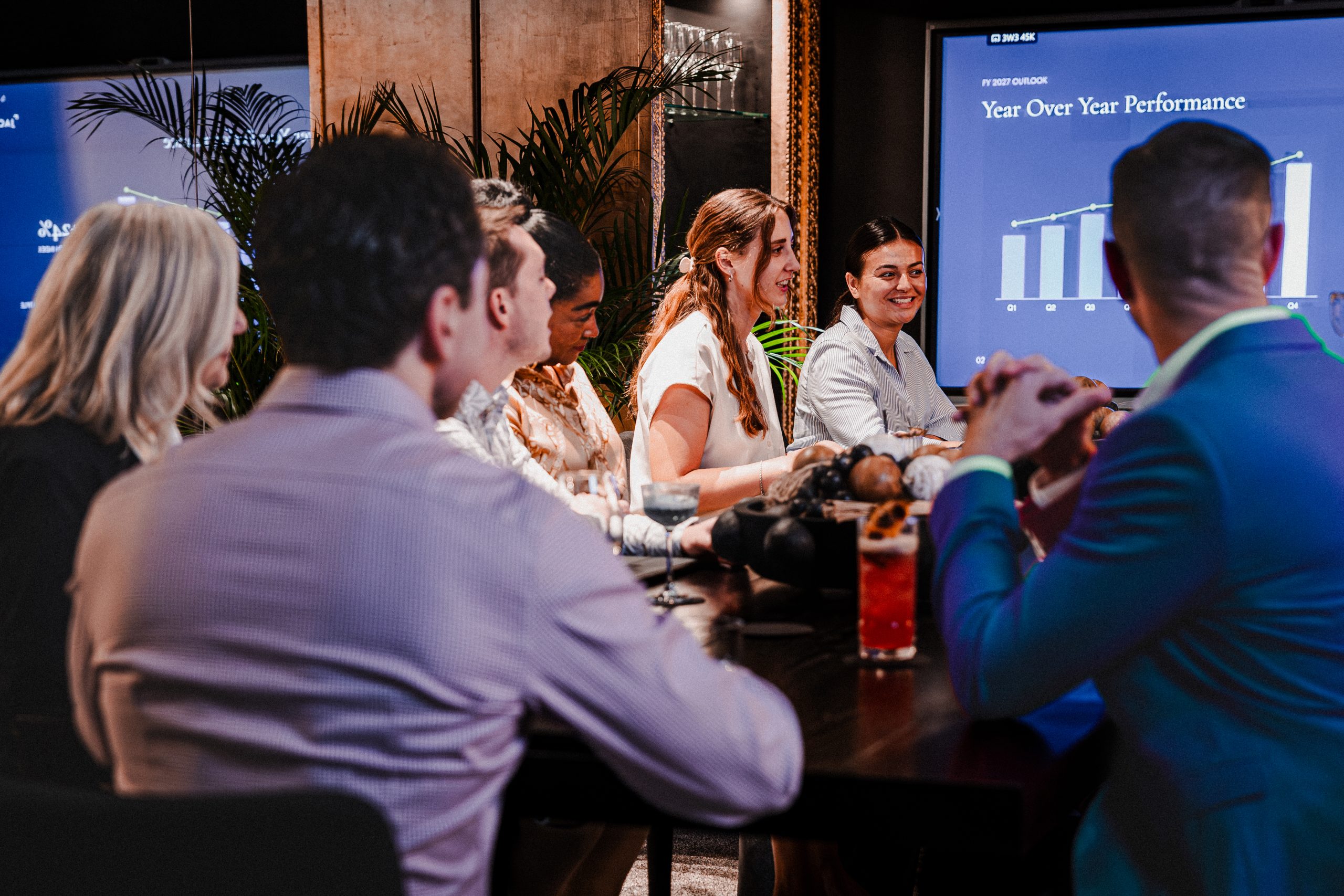 Team in the Boardroom watches a year-over-year performance chart on a large screen under a crystal chandelier