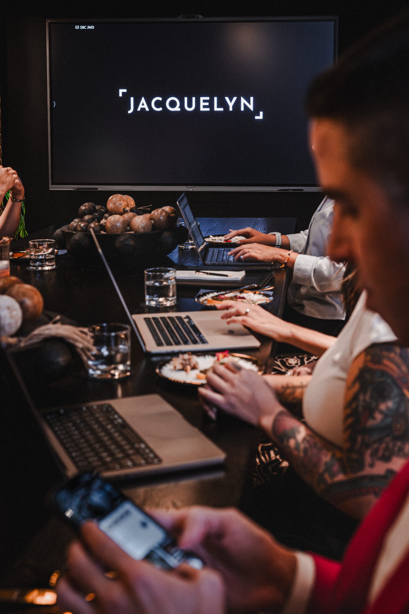 People work on laptops around the Boardroom table, with a screen showing the Jacquelyn logo