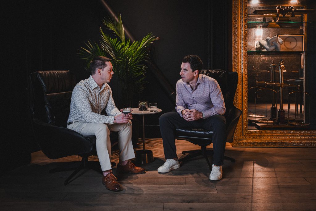 Two men talk in leather chairs in a private corner of the Boardroom, beside a gilded display case