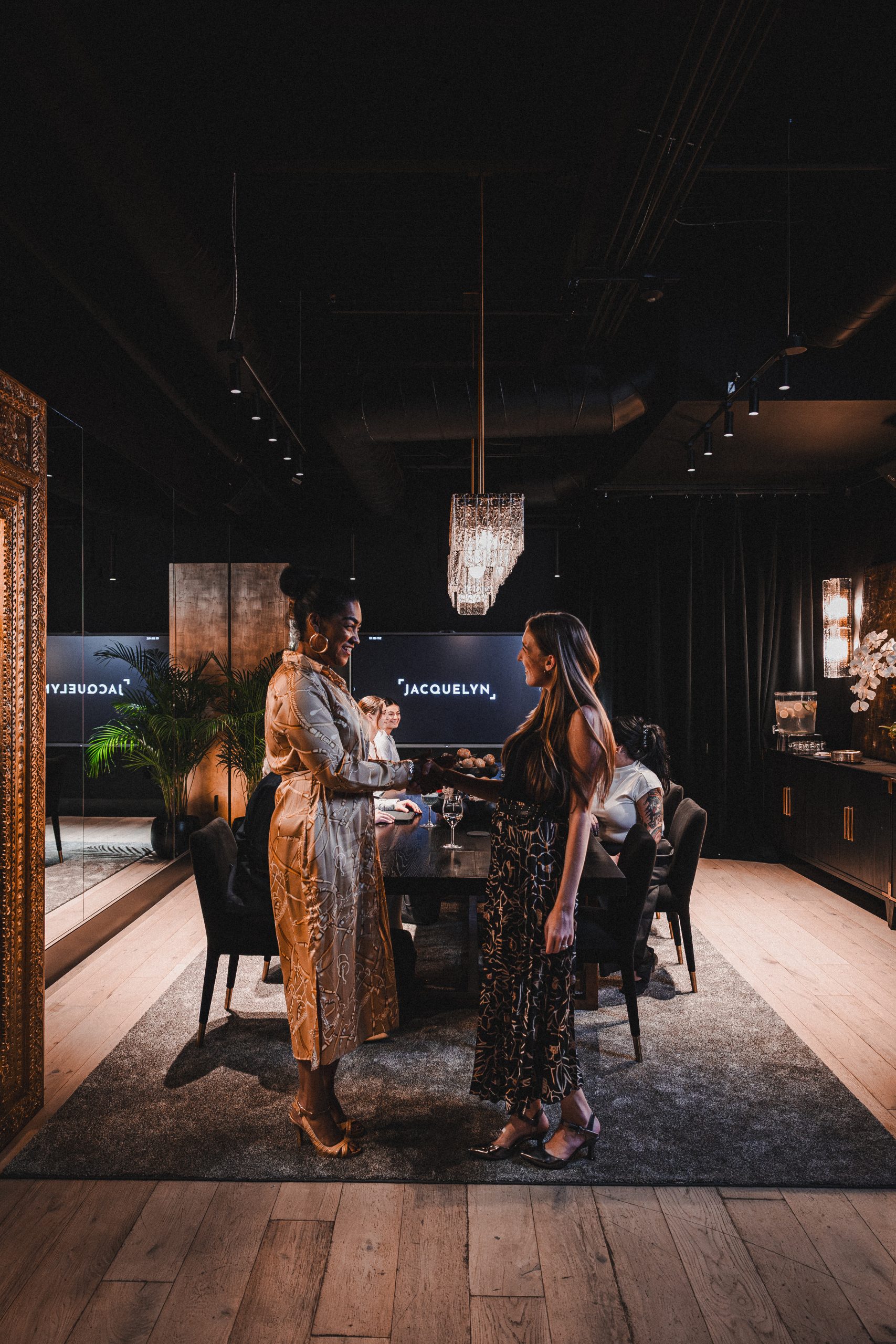 Two women greet each other in the Boardroom as others sit at the conference table behind them